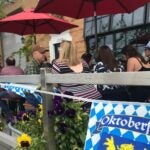 Oktoberfest revelers sit at picnic tables outside Dorchester Brewing Company in Boston. The patio railing has an Oktoberfest banner visible, as well as leafy, bright yellow violets growing in abundance.