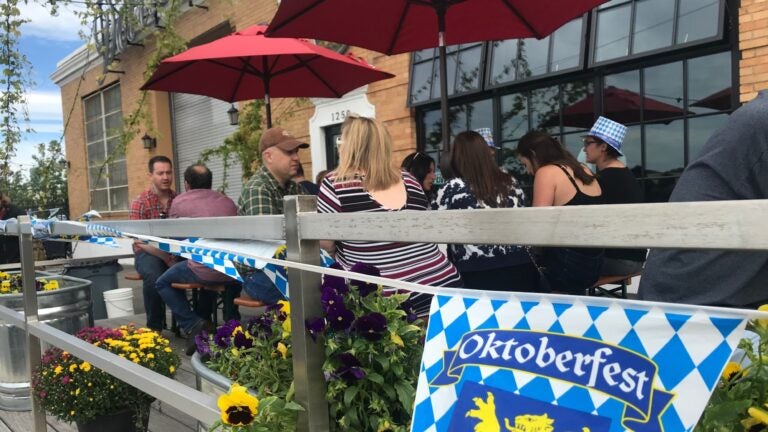 Oktoberfest revelers sit at picnic tables outside Dorchester Brewing Company in Boston. The patio railing has an Oktoberfest banner visible, as well as leafy, bright yellow violets growing in abundance.