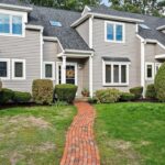The outside of a town home with gray clapboard siding. It's at the end of a brick walkway that bisects a green lawn.