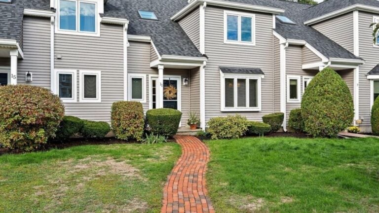 The outside of a town home with gray clapboard siding. It's at the end of a brick walkway that bisects a green lawn.