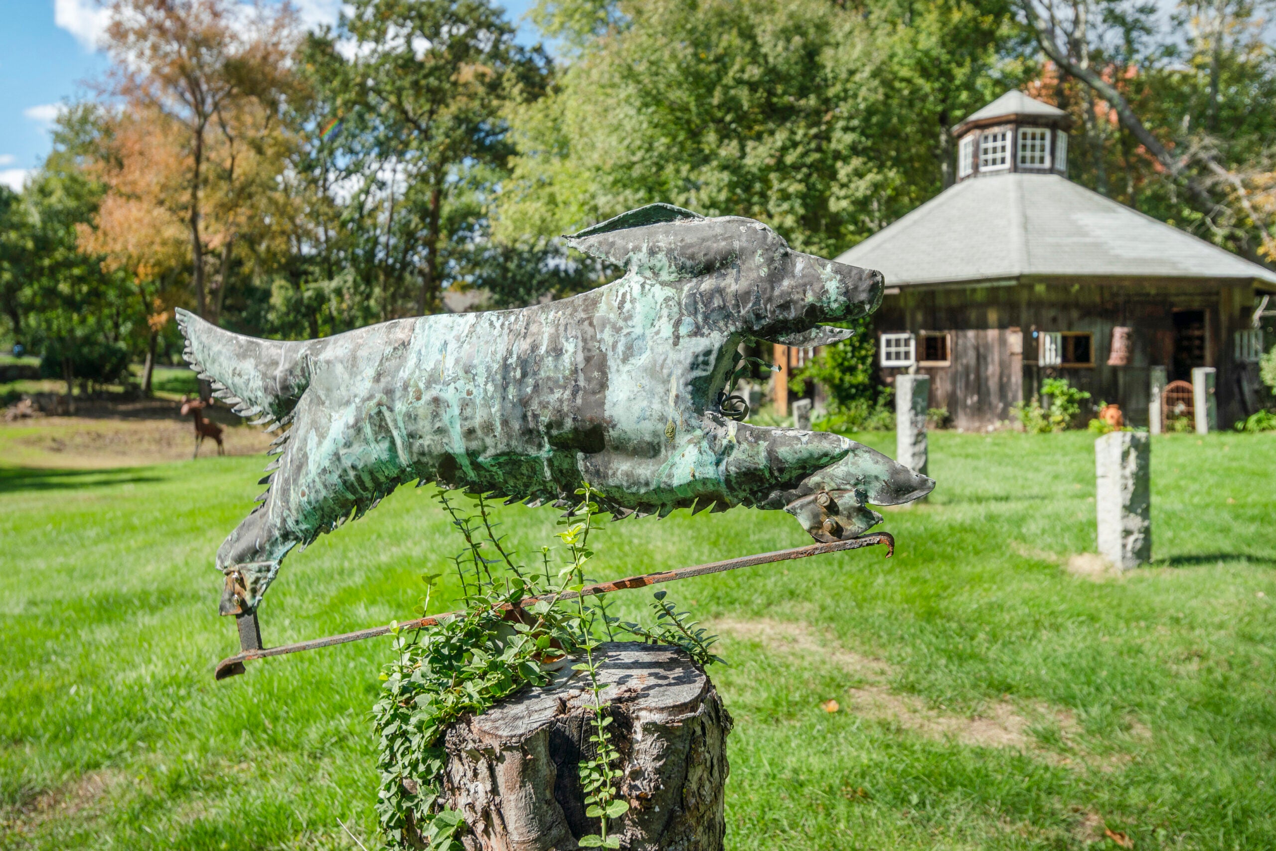 A copper statue shaped like a running dog on a tree stump in front of an octagonal barn.