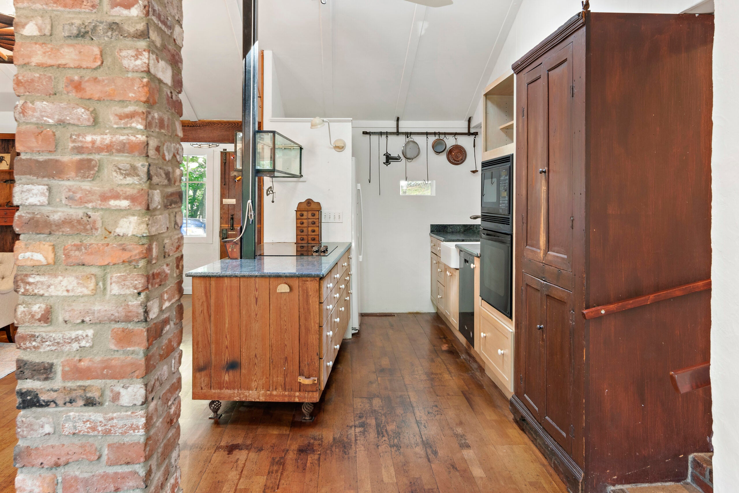 A brick column, a natural wood kitchen, white walls, and a pot rack.