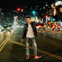 Comedian Dane Cook, wearing a leather jacket, poses in the street with a microphone stand.