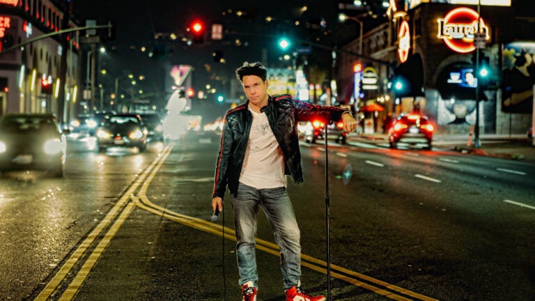 Comedian Dane Cook, wearing a leather jacket, poses in the street with a microphone stand.