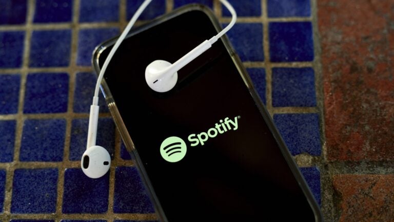 alt = the Spotify logo shown on the screen of a smartphone with white earbuds draped over the phone