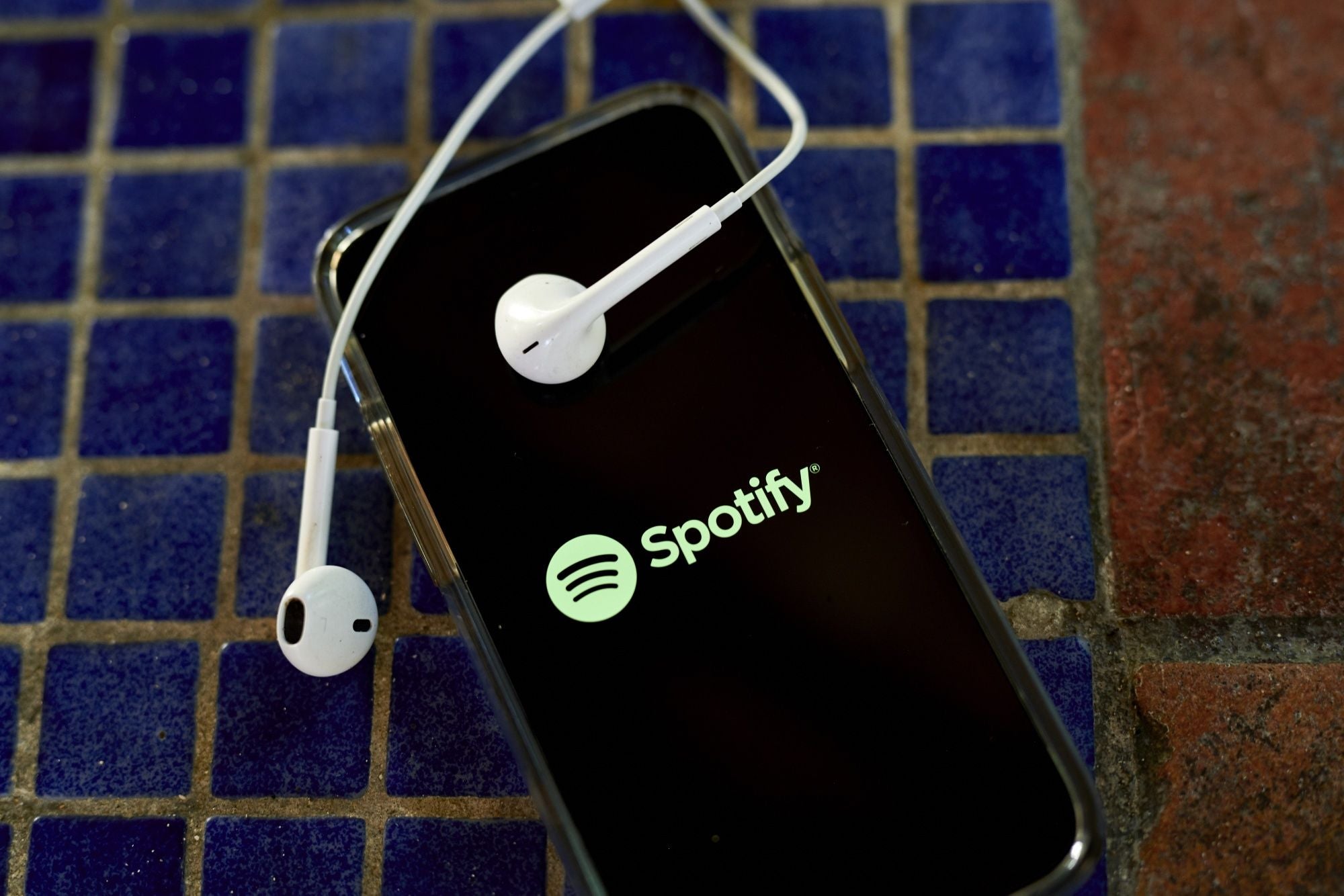 alt = the Spotify logo shown on the screen of a smartphone with white earbuds draped over the phone