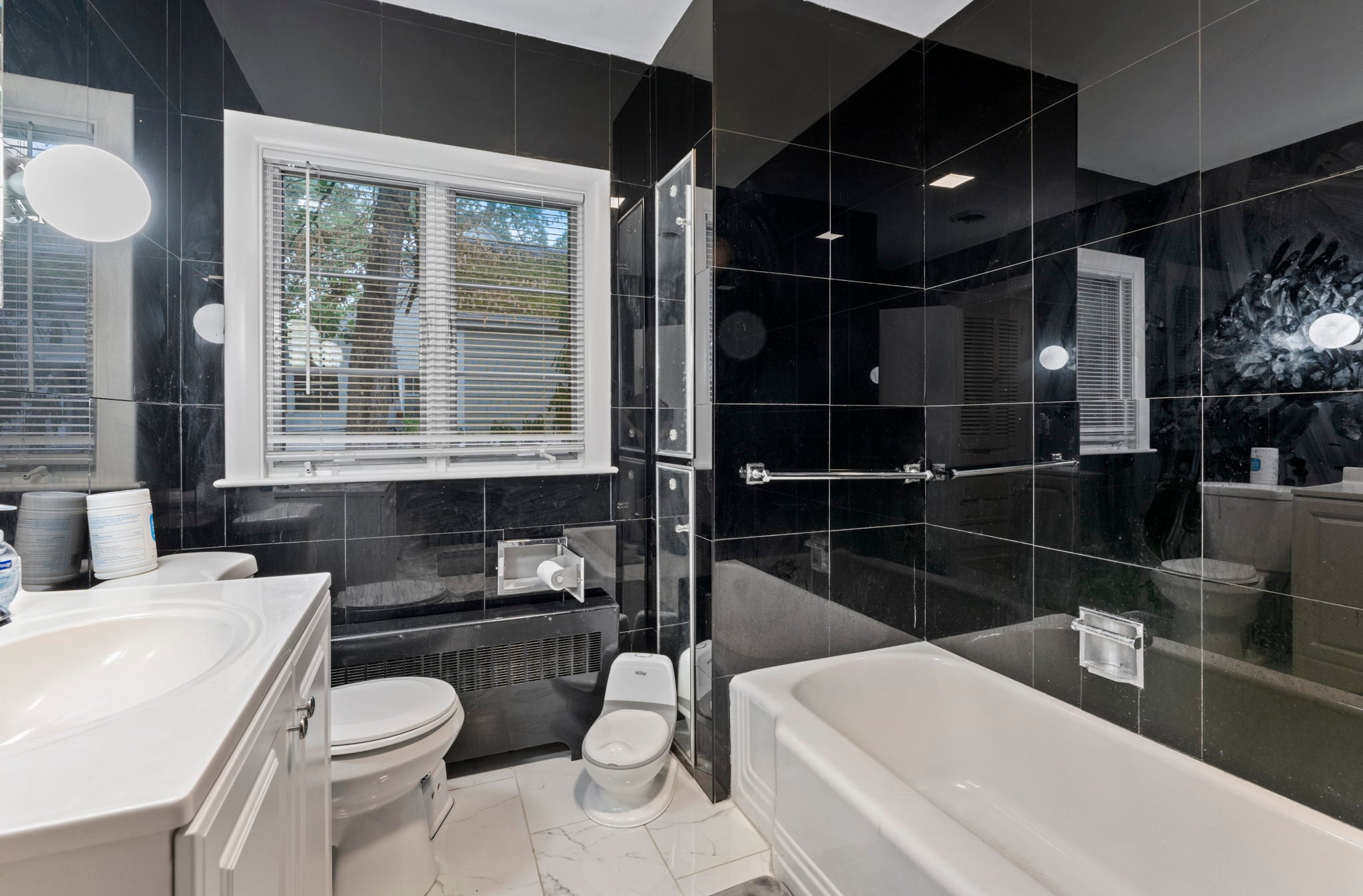 A bathroom with a tub/shower combination, a white vanity, a white tile floor, and large-format black tile walls.
