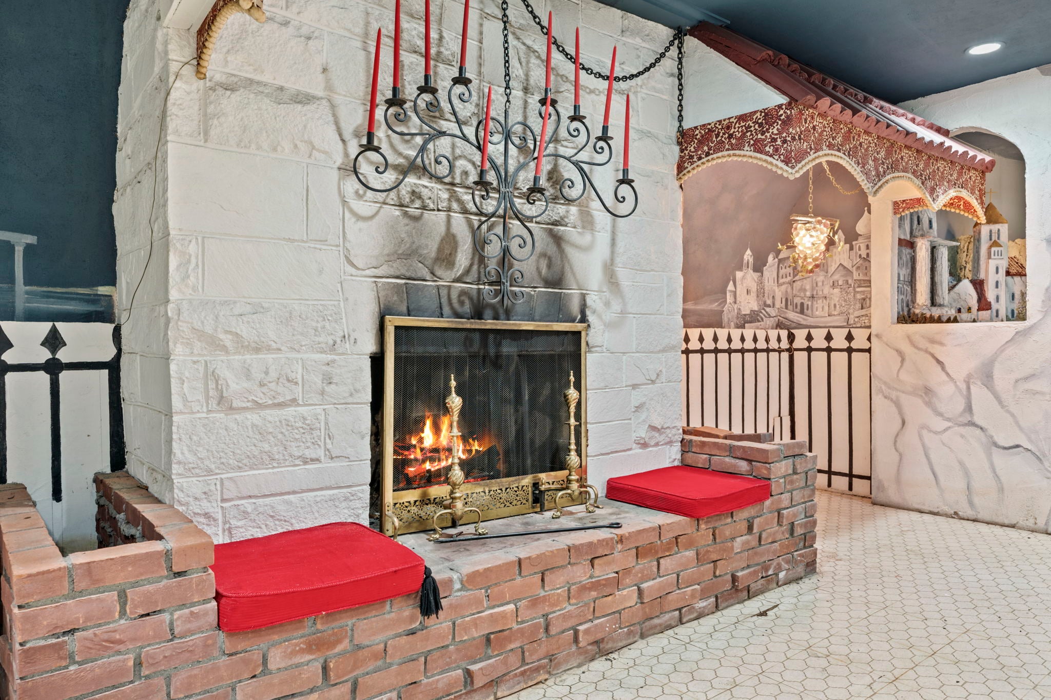 A candelabra with red candles is mounted above a working fireplace with a brick hearth and two red seat cushions.