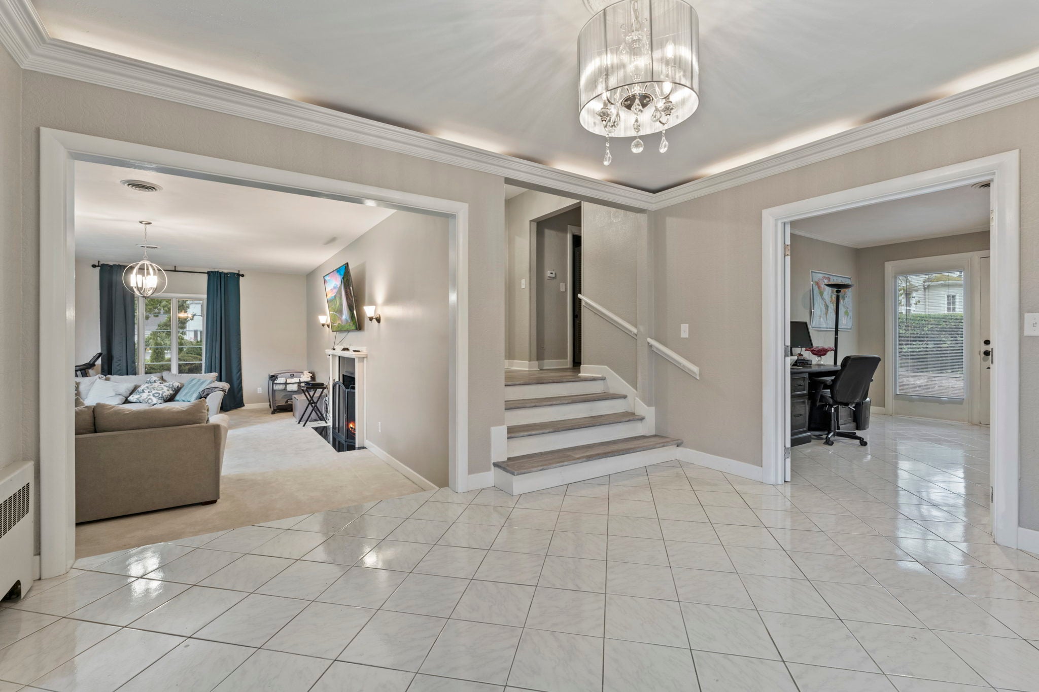 A circular chandelier hangs over a foyer with a white tile floor and mounting above the crown molding.