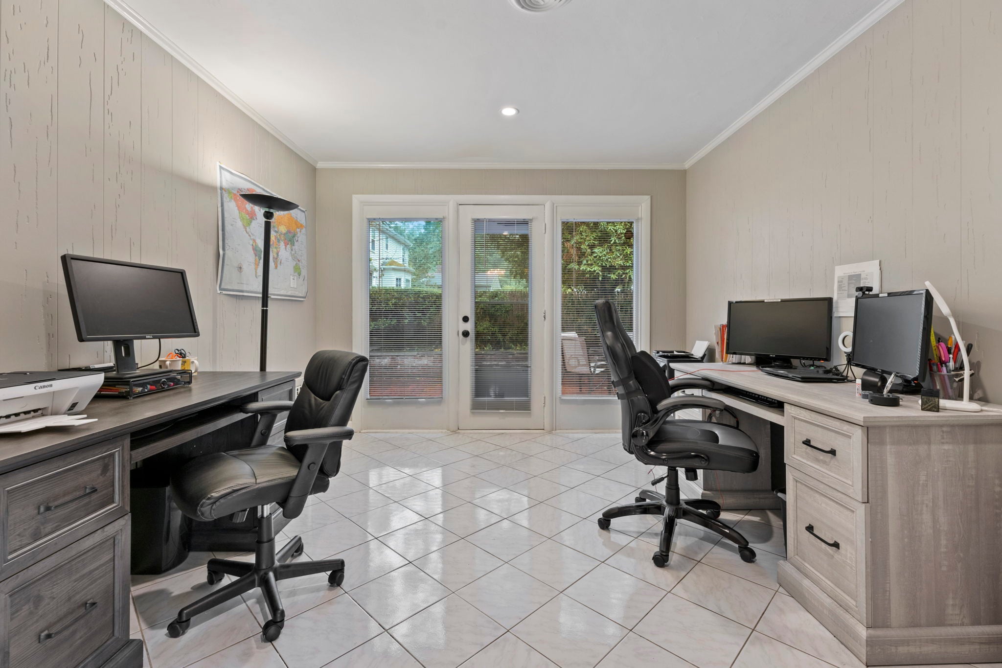 An office with two desks, wood paneling, a single door to a patio, and a white tile floor.