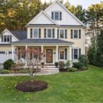 The exterior of a three-story yellow and white house with a two-car garage, a green lawn, mature landscaping, green shutters, and a farmer's porch.