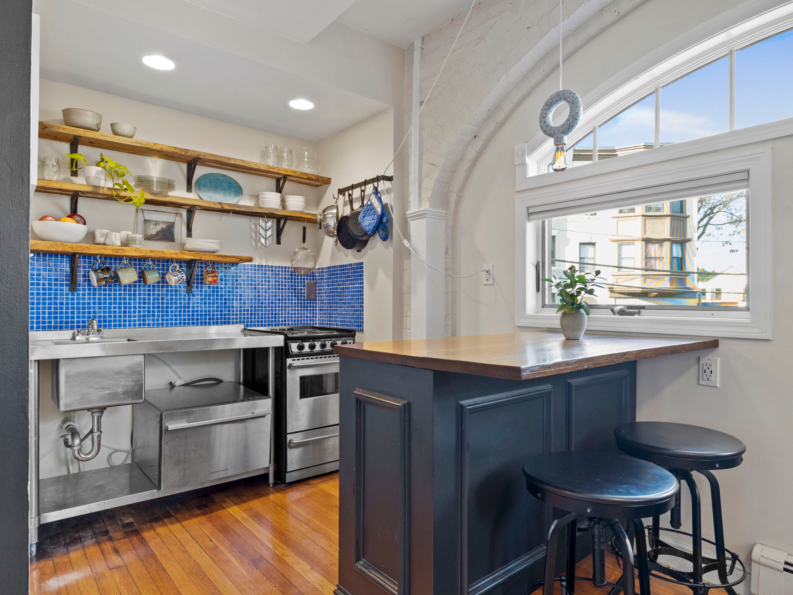 A kitchen with a bright blue mosaic backsplash, open shelving, exposed brick, and a peninsula with seating for two.