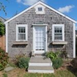 A cedar shake cottage with two window boxes. The trees are green and the sky is sunny.