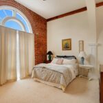 A bedroom with exposed brick, carpeting, a Palladian window, and a bed with two night stands.