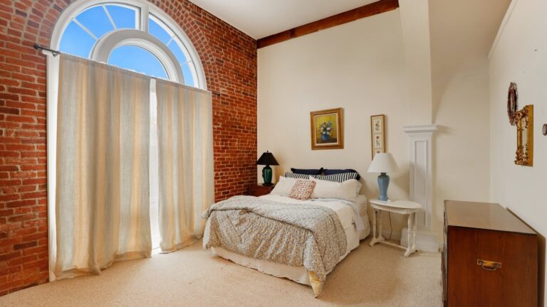 A bedroom with exposed brick, carpeting, a Palladian window, and a bed with two night stands.