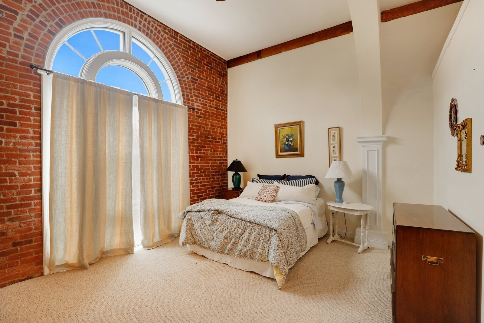 A bedroom with exposed brick, carpeting, a Palladian window, and a bed with two night stands.