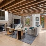 A living room with a myriad of exposed beams, exposed brick, shiplap, a fireplace, light-colored, wide-plank wood flooring, and double doors to a balcony.