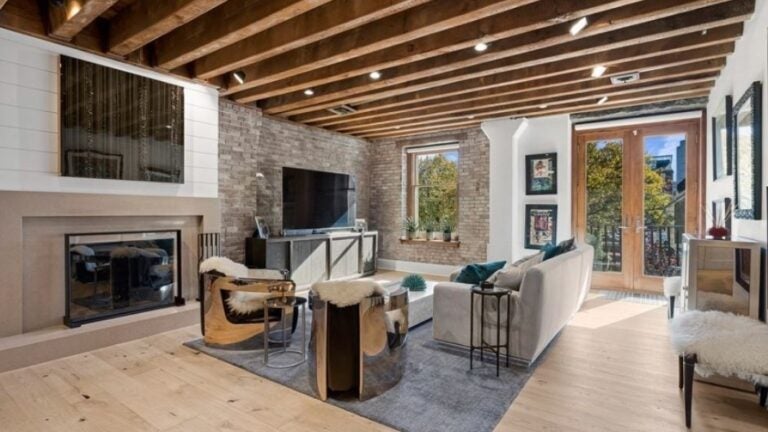 A living room with a myriad of exposed beams, exposed brick, shiplap, a fireplace, light-colored, wide-plank wood flooring, and double doors to a balcony.