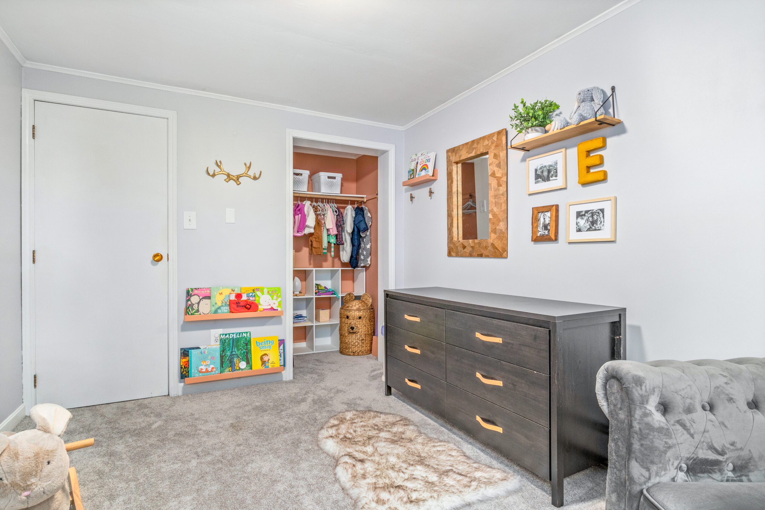 A bedroom with gray carpeting, a orange closet without a door, toddler clothes and toys, and a six-drawer dresser.