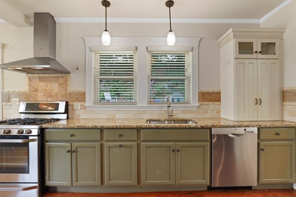 View of a kitchen with white walls, two windows, green cabinetry, and two pendant lights.
