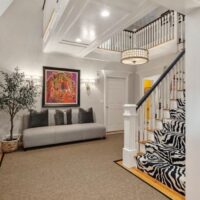 A carpeted foyer with a gray couch straight ahead, griege walls, a drum shade light, and stairs carpeted in a zebra print.