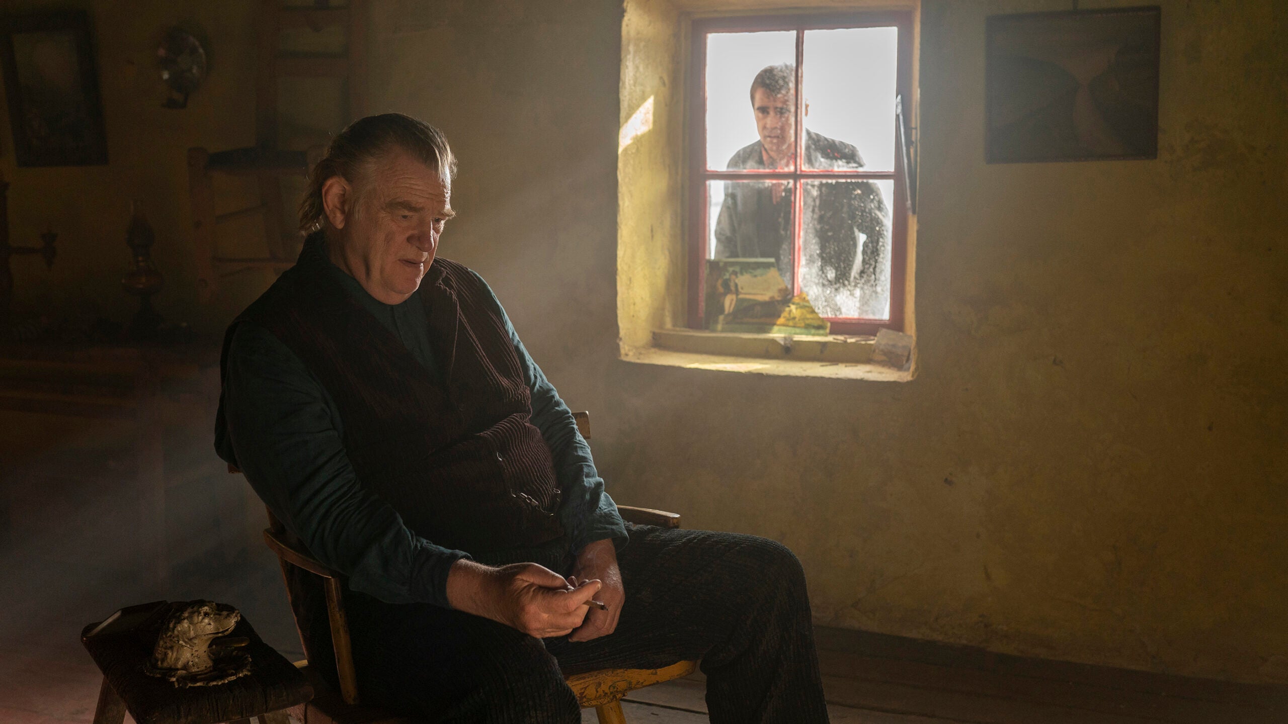 Brendan Gleeson sitting in the foreground and Colin Farrell looking at him through a window in "The Banshees of Inisherin."