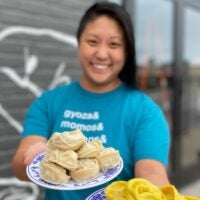 Mei Mei founder and chef Irene Li greets the camera outside the South Boston factory with two plates of dumplings, in focus.