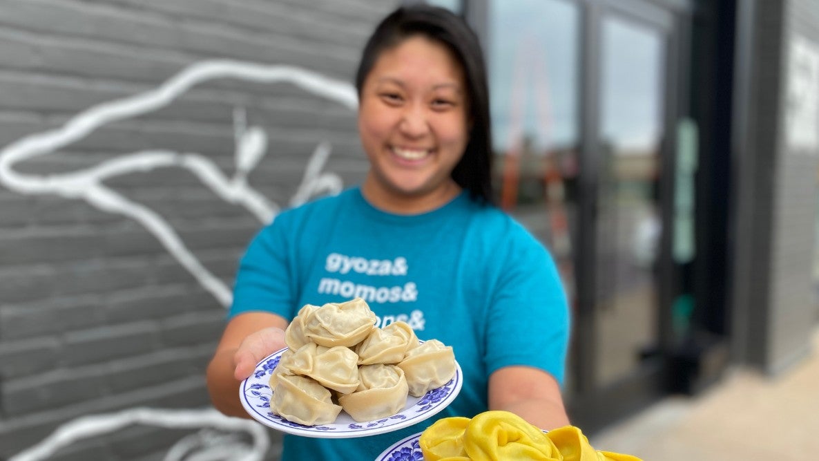 Mei Mei founder and chef Irene Li greets the camera outside the South Boston factory with two plates of dumplings, in focus.