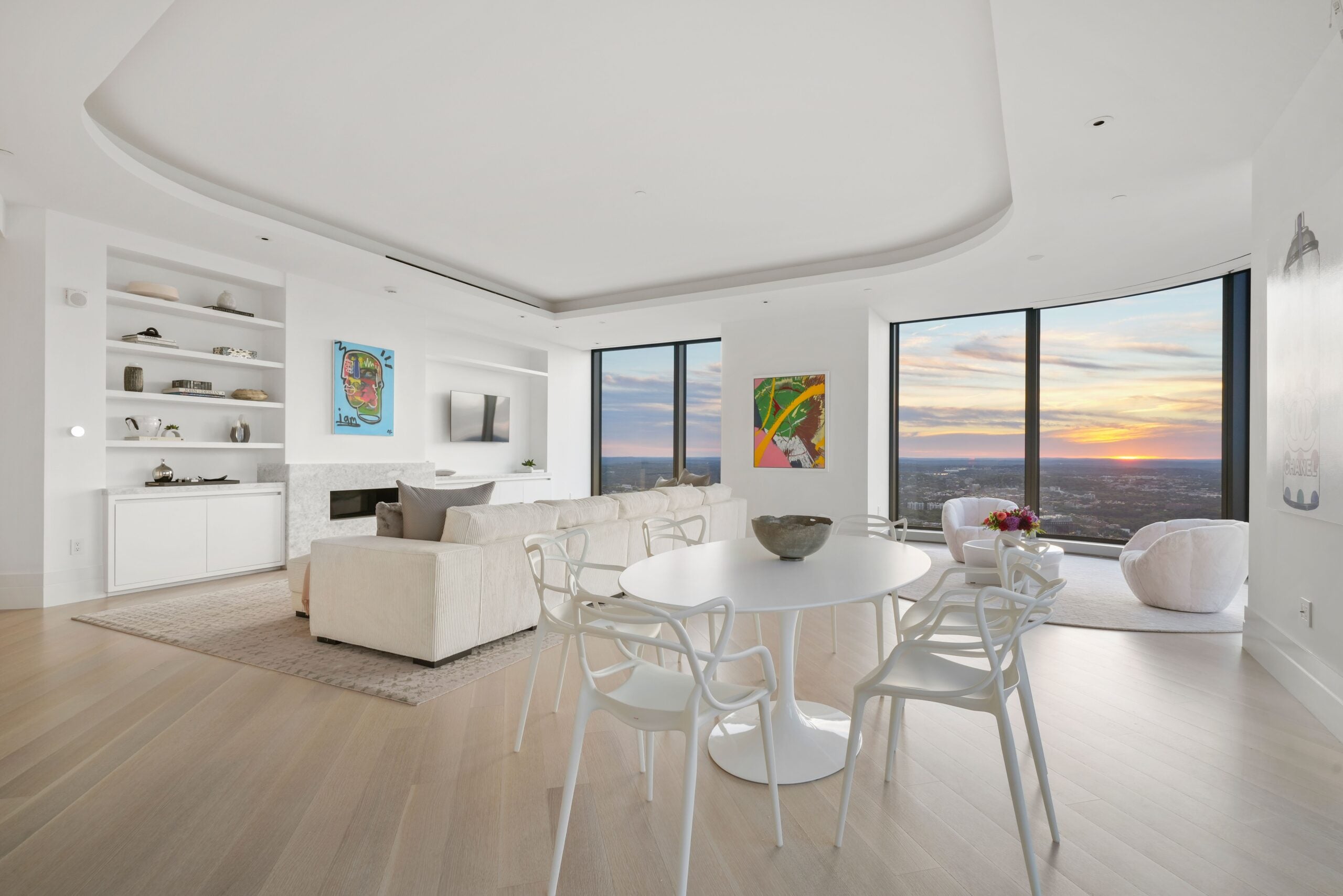 One Dalton living room with couch, seating area, fireplace, casual eating area and sweeping views of the city. Sun just above the horizon in the distance. Hardwood floors in space.