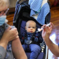 George O’Connor, age 1 from East Boston, watches from his baby carriage as a COVID vaccine is given to his grandmother, Josie O’Connor (left) during a vaccination clinic where flu shots were also being offered.