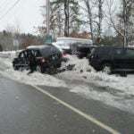alt = a damaged black Jeep Compass pictured after a crash on a snowbank, next a green sign that reads "Welcome to Plaistow Village. Slow down." and other parked cars with visible damage. The crash took place in the area of Route 125 and Route 121A in Plaistow, New Hampshire.