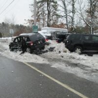 alt = a damaged black Jeep Compass pictured after a crash on a snowbank, next a green sign that reads "Welcome to Plaistow Village. Slow down." and other parked cars with visible damage. The crash took place in the area of Route 125 and Route 121A in Plaistow, New Hampshire.