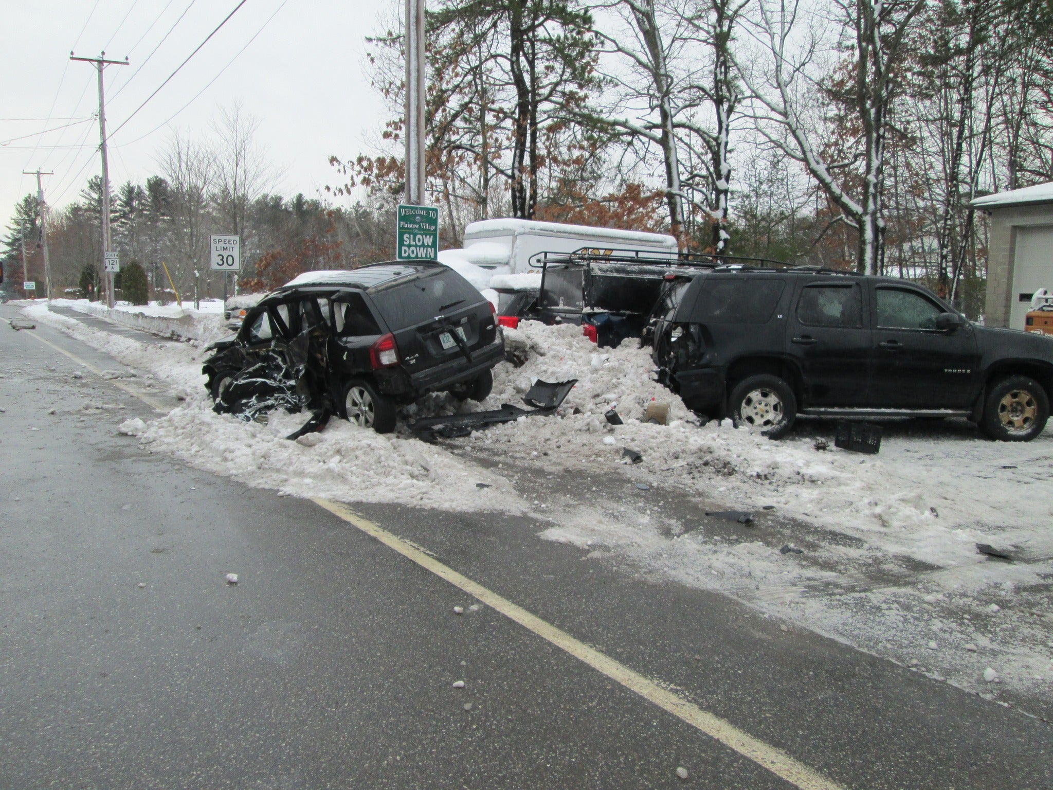 alt = a damaged black Jeep Compass pictured after a crash on a snowbank, next a green sign that reads "Welcome to Plaistow Village. Slow down." and other parked cars with visible damage. The crash took place in the area of Route 125 and Route 121A in Plaistow, New Hampshire.