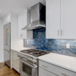 A kitchen with white walls, cabinets, and counters, and a swirly blue tile backsplash to consider for your next home improvement project.