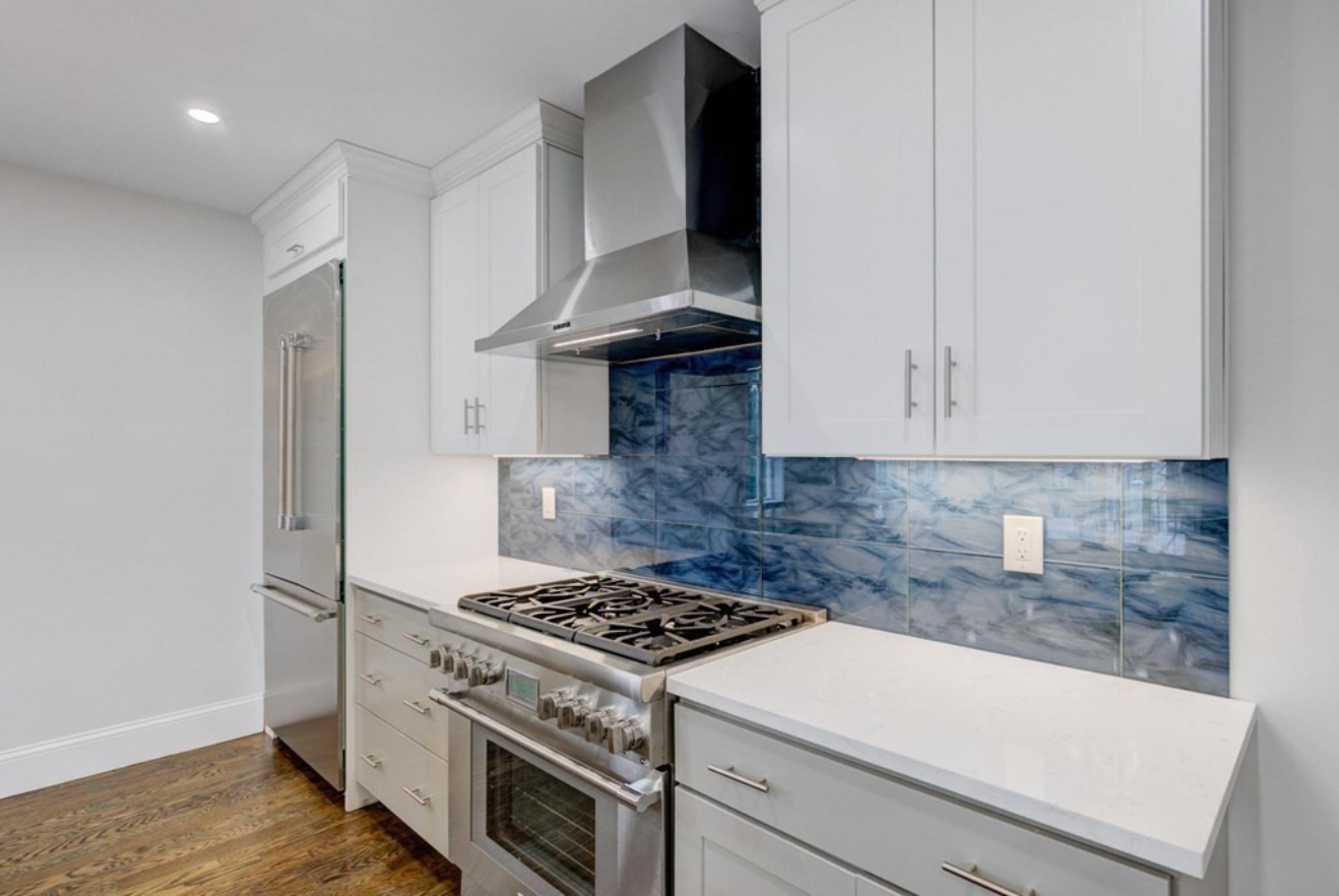 A kitchen with white walls, cabinets, and counters, and a swirly blue tile backsplash to consider for your next home improvement project.