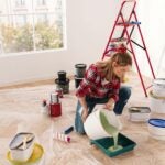 Young Caucasian woman alone in new apartment with big window pouring green paint preparing for painting walls with roller brush. Home repair and renovation. Redesigning room