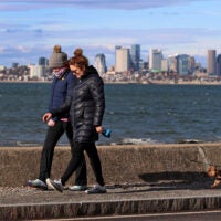 People go for a brisk winter walk with a dog along a breakwater in Squantum with Boston in the background.