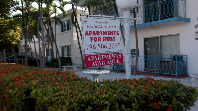 alt = A white sign with red lettering reading 'Apartments for Rent' hangs in front of a building surrounded by bushes in Miami Beach, Florida.