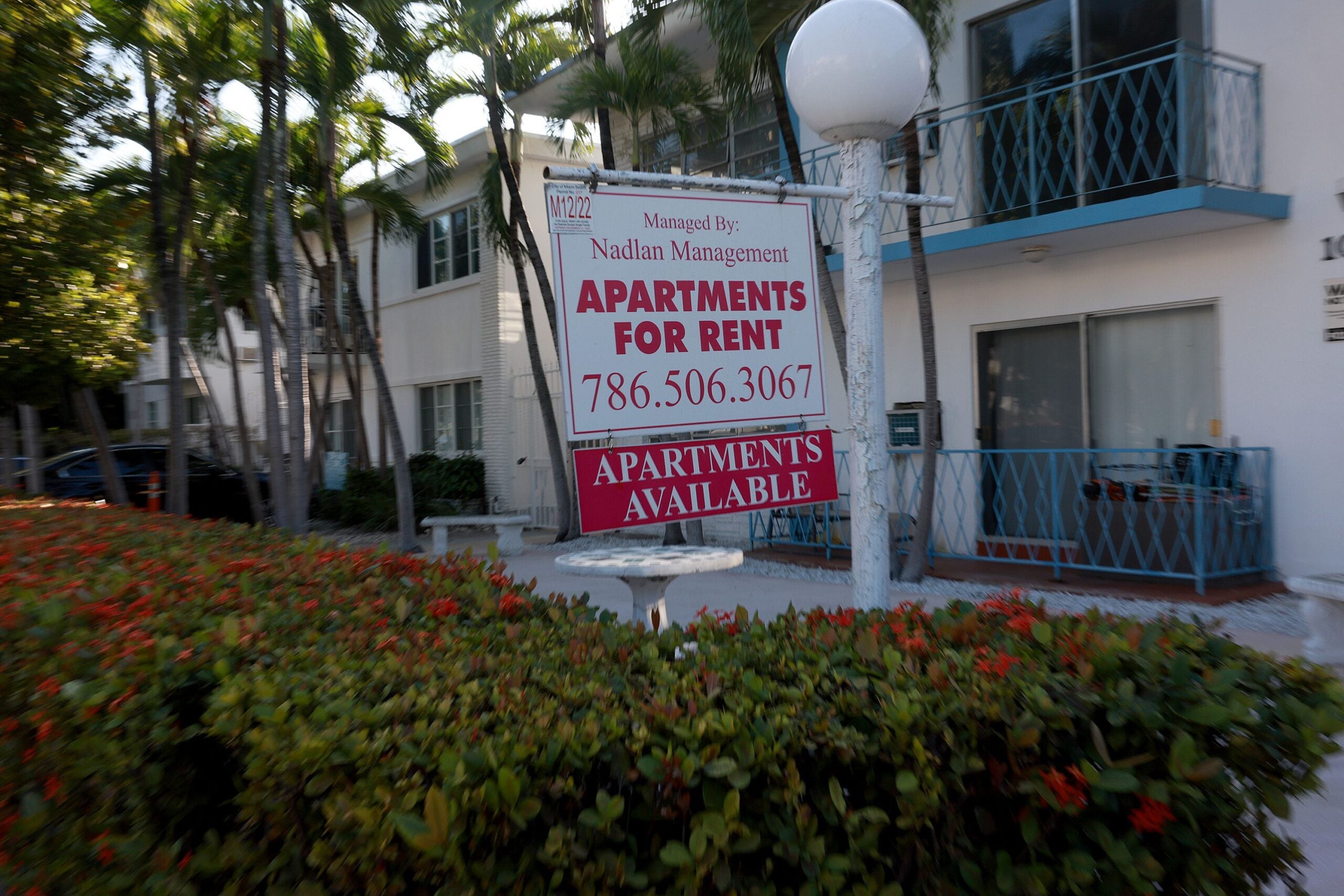alt = A white sign with red lettering reading 'Apartments for Rent' hangs in front of a building surrounded by bushes in Miami Beach, Florida.
