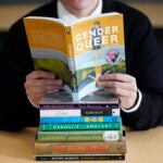 alt = Amanda Darrow, director of youth, family and education programs at the Utah Pride Center, poses with books that have been the subject of complaints from parents in Salt Lake City on Dec. 16, 2021.