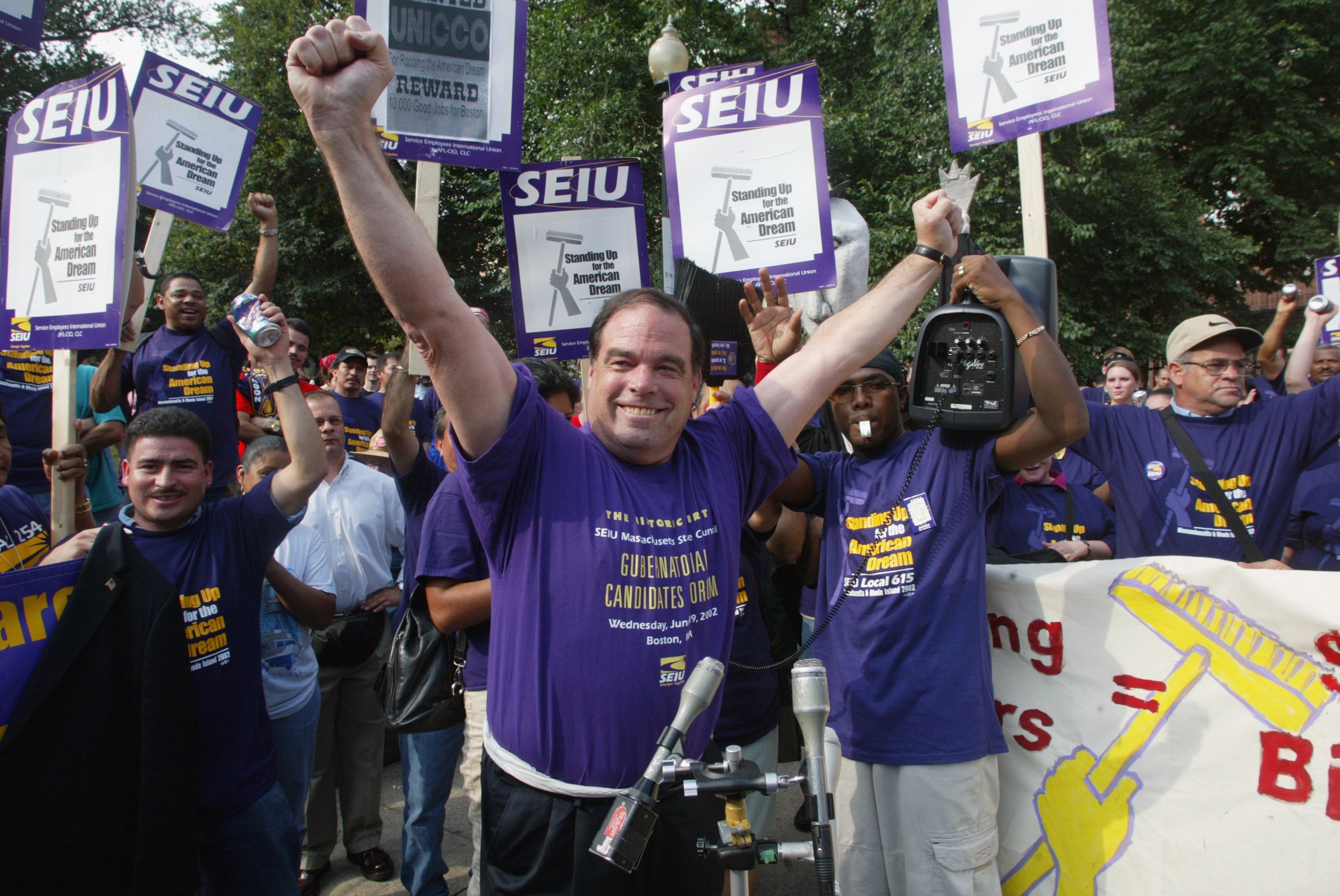 alt = Senate President Thomas Birmingham, wearing a purple SEIU t-shirt, lifts his arms in the air at a rally in support of janitors at the Park Street T stop on the Boston Common.