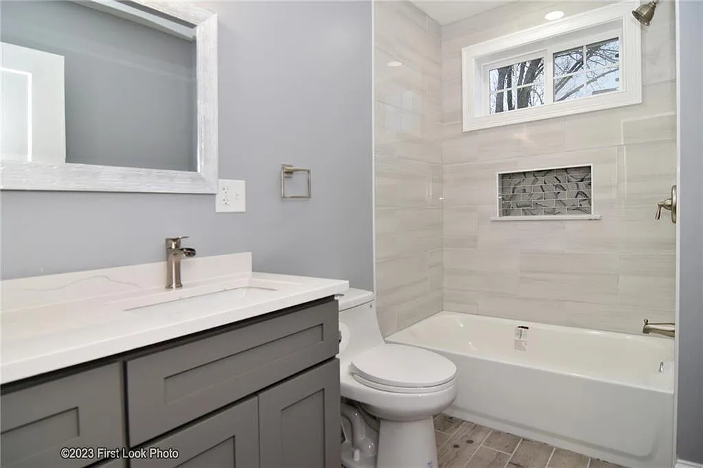 A bathroom with gray walls, a white shower/bathtub, light gray tiling and a double-hung window.