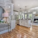 A home with gray and white walls, white kitchen cabinetry, a circular gold mirror, pendant lights, and a fireplace. The floor is wood.