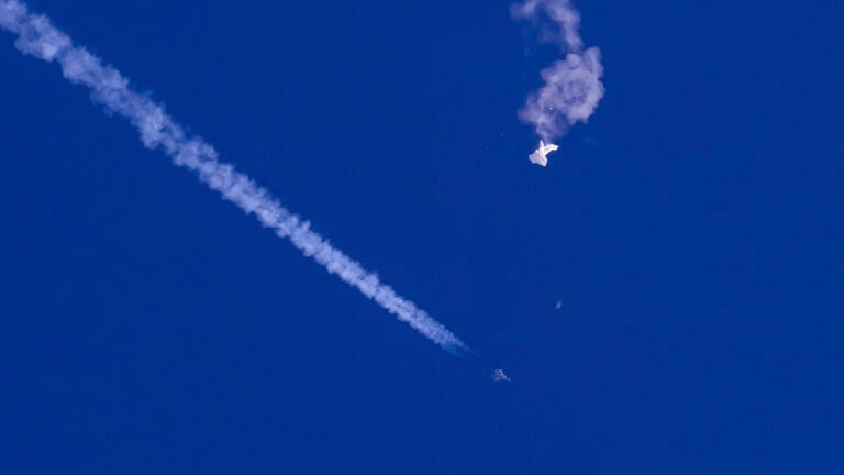 In this photo provided by Chad Fish, the remnants of a large balloon drift above the Atlantic Ocean, just off the coast of South Carolina, with a fighter jet and its contrail seen below it, Saturday, Feb. 4, 2023.