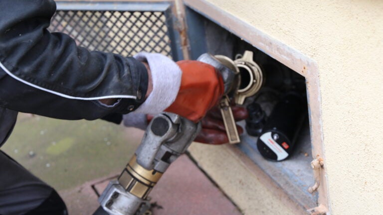 A person wearing red gloves puts a nozzle into a receptor for home heating oil on the side of a home.