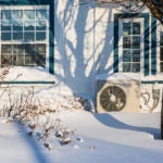 Heat pump unit on the side of a white home with blue trim. It is winter.