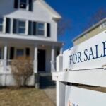 A white "for sale" sign in front of a white Colonial with black shutters and a farmer's porch. The grass is winter brown.