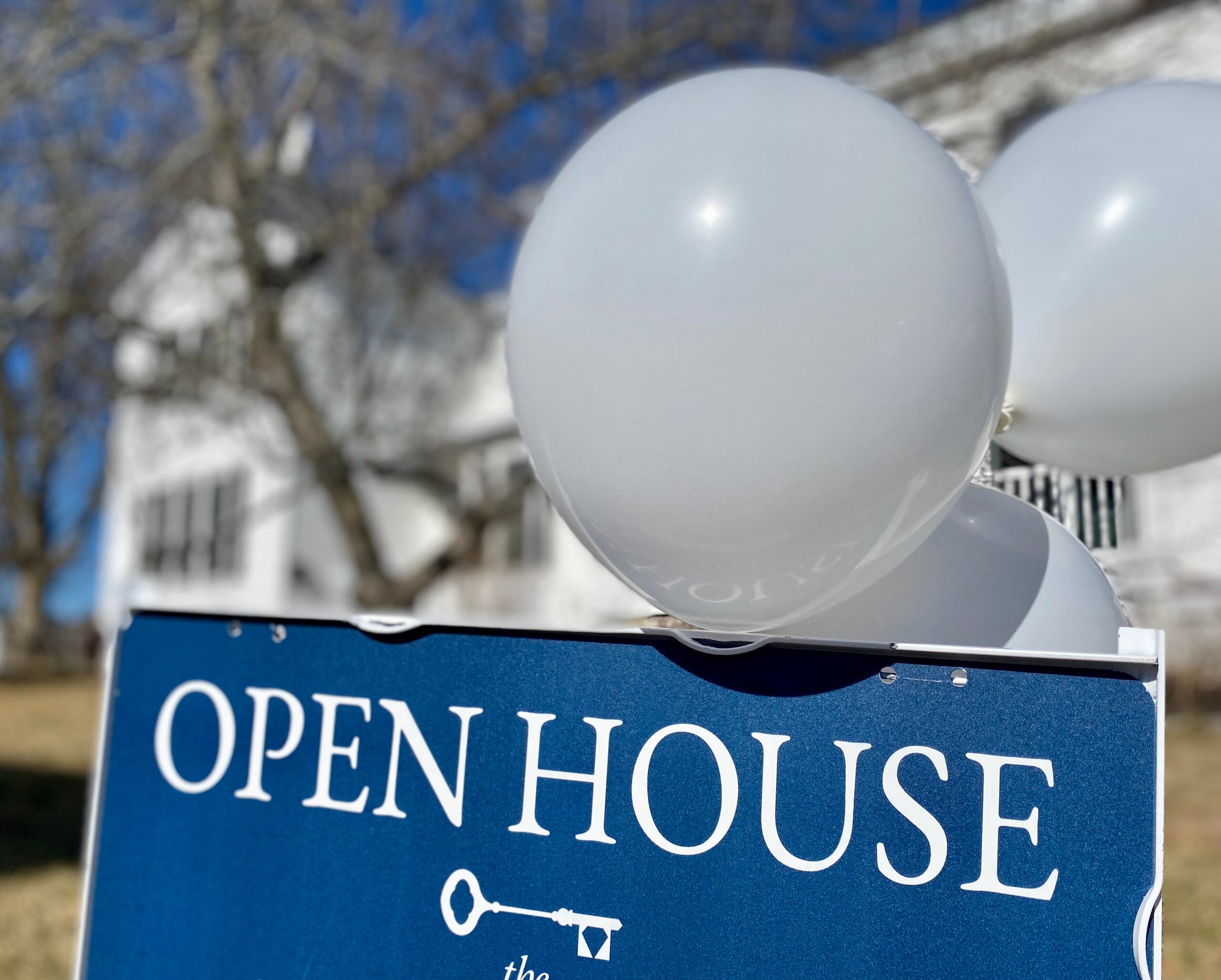 An open house sign with white lettering on a blue background. Balloons float overhead.