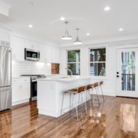A kitchen with white walls and white Shaker-style cabinets, stainless steel appliances, double-hung windows, hardwood floors, and exposed brick accents.