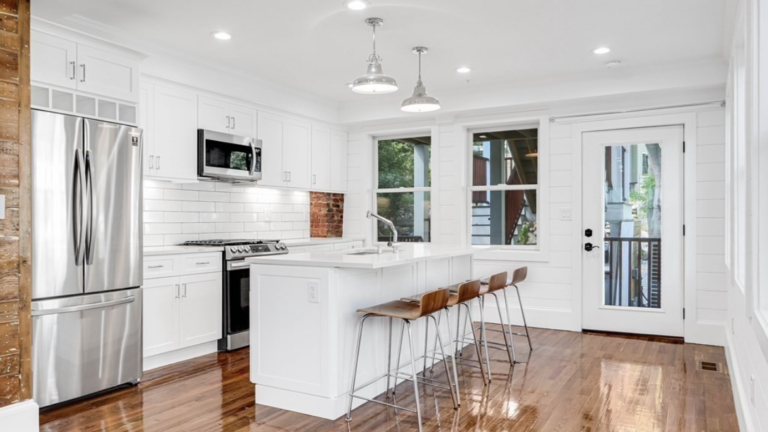 A kitchen with white walls and white Shaker-style cabinets, stainless steel appliances, double-hung windows, hardwood floors, and exposed brick accents.